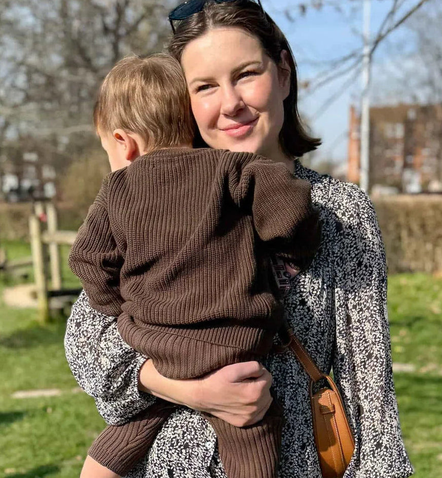 Woman holding a child in a park with trees and buildings in the background